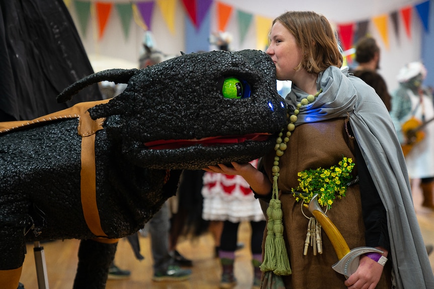 A girl dressed in a grey cape and a brown garment with yellow flowers kisses the head of a black robotic dragon with green eyes.