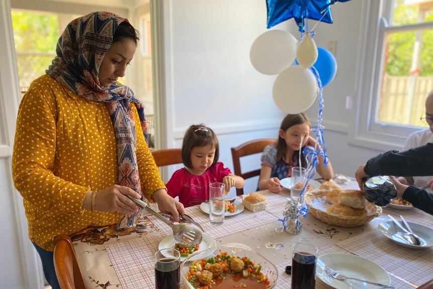 A woman fills a plate with food. Next to her two young girls are seated at a table with plates of food in front of them