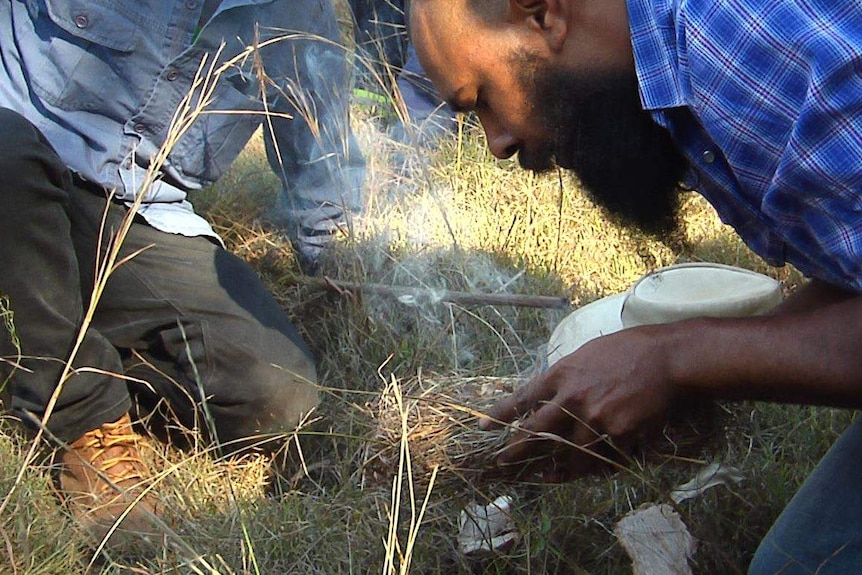 An Aboriginal man leans over a handful of smoking grass to fan flames.