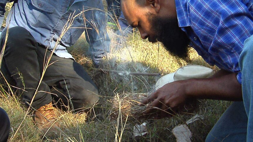 An Aboriginal man leans over a handful of smoking grass to fan flames