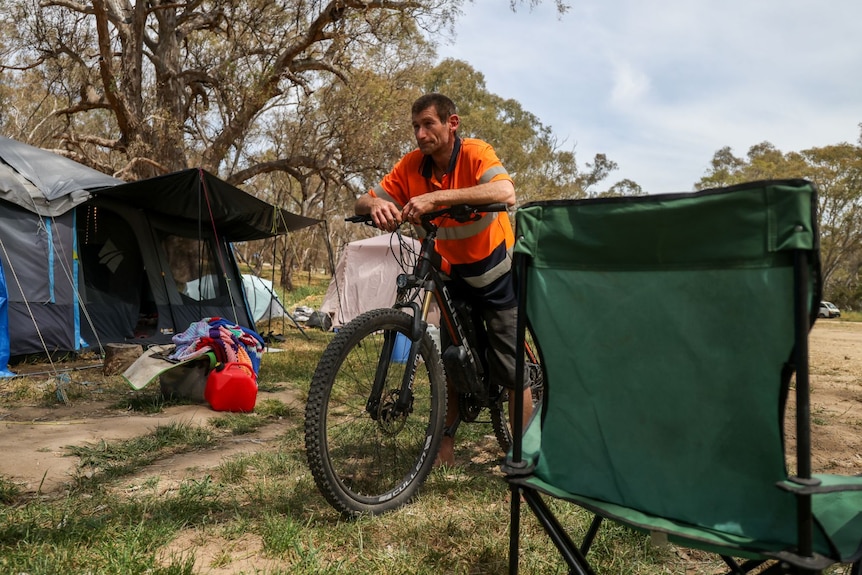A man standing with his bike outside a tent and a camping chair.