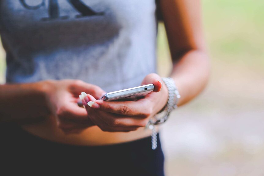 Close-up of a woman holding her smart phone. She wears a grey t-shirt and silver bracelets.