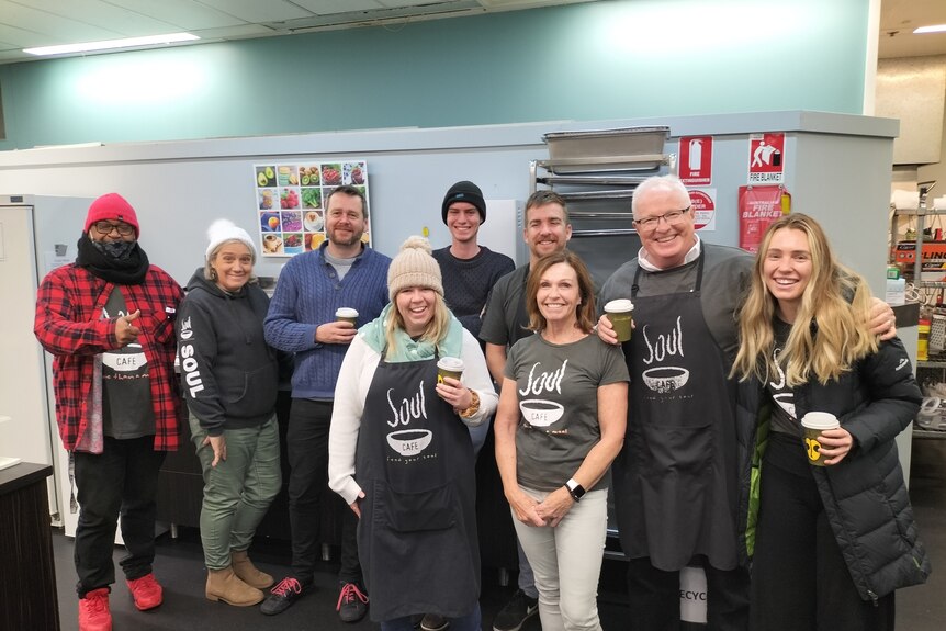 A group of nine men and women, most wearing matching aprons, stand smiling in a soup kitchen.