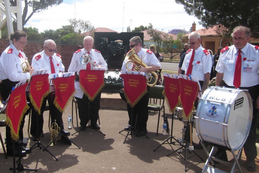 Seven members of the brass band are performing outside at Whyalla wearing long-sleeve shirts and black pants.