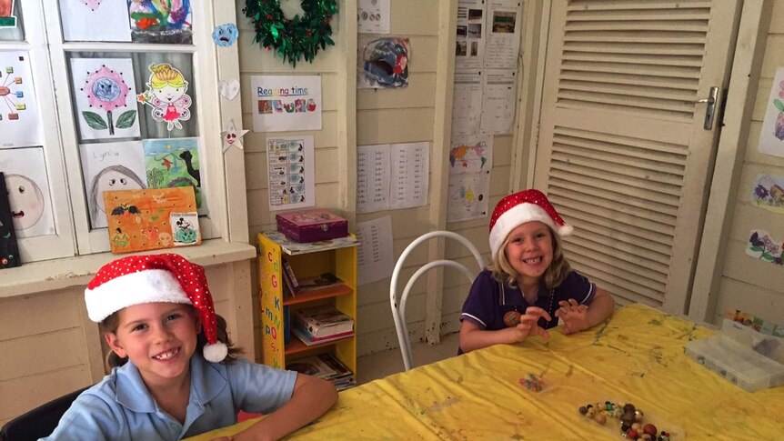 Two distance education students wearing Santa hats sit at a table in a makeshift classroom with craft supplies.