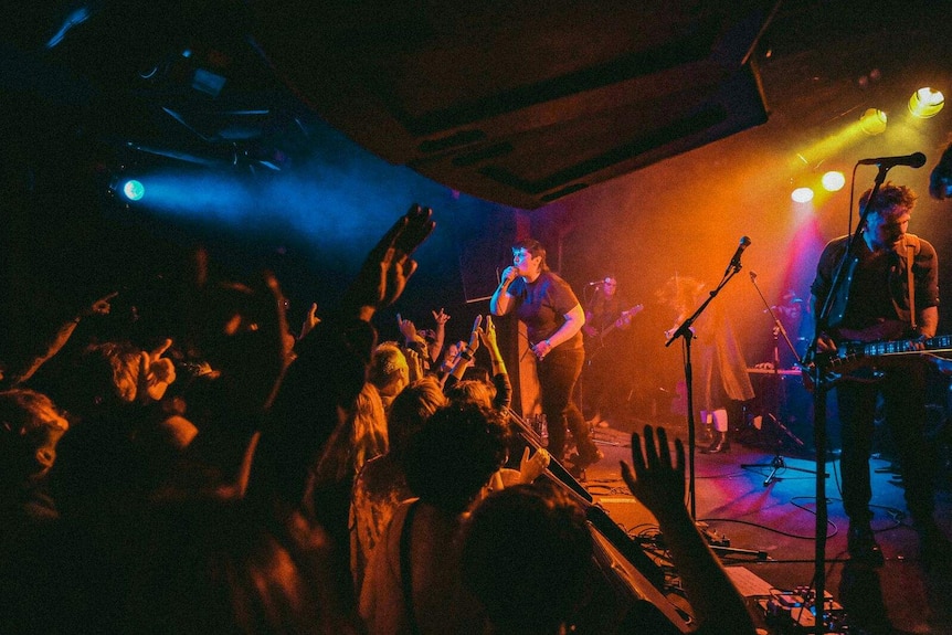 A woman sings into a microphone on stage with a crowd waving arms and a yellow light over the stage.