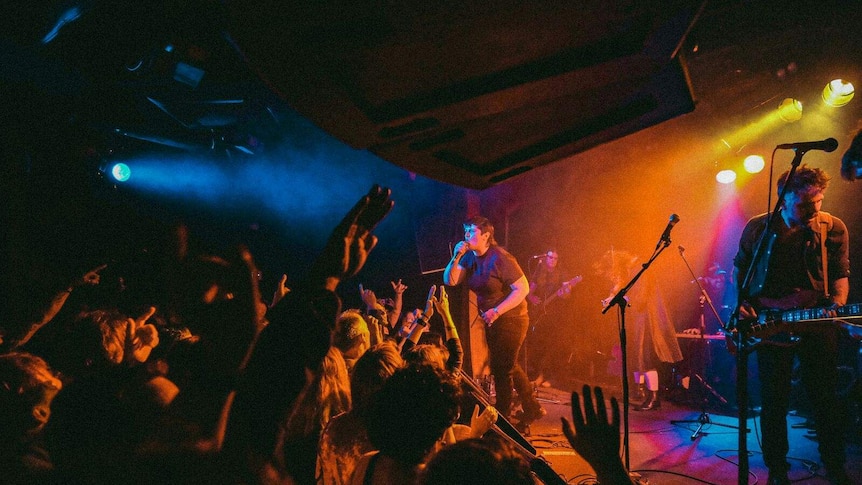 A woman sings into a microphone on stage with a crowd waving arms and a yellow light over the stage.