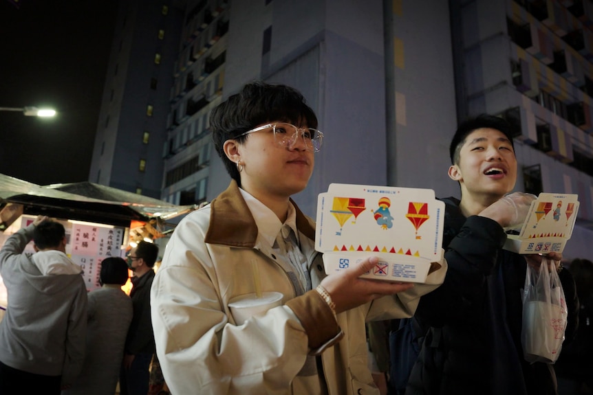 Two young people stand outside in a night market eating out of takeaway boxes