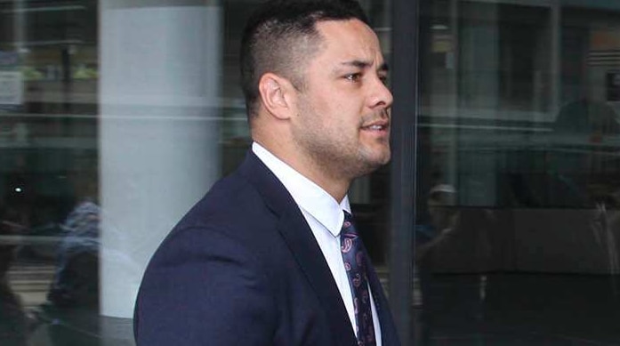 A young man in a dark suit walks past a large glass window. A woman in dark sunglasses walks behind him.