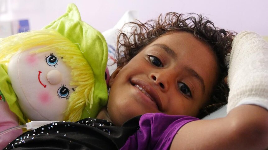 A smiling child with a bandaged arm and a colourful doll in a hospital bed