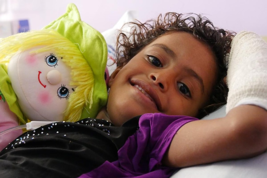 A smiling child with a bandaged arm and a colourful doll in a hospital bed