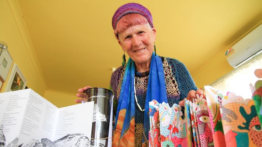 A woman wearing colourful clothes smiles down on the camera which is focussed on the art works before her. 