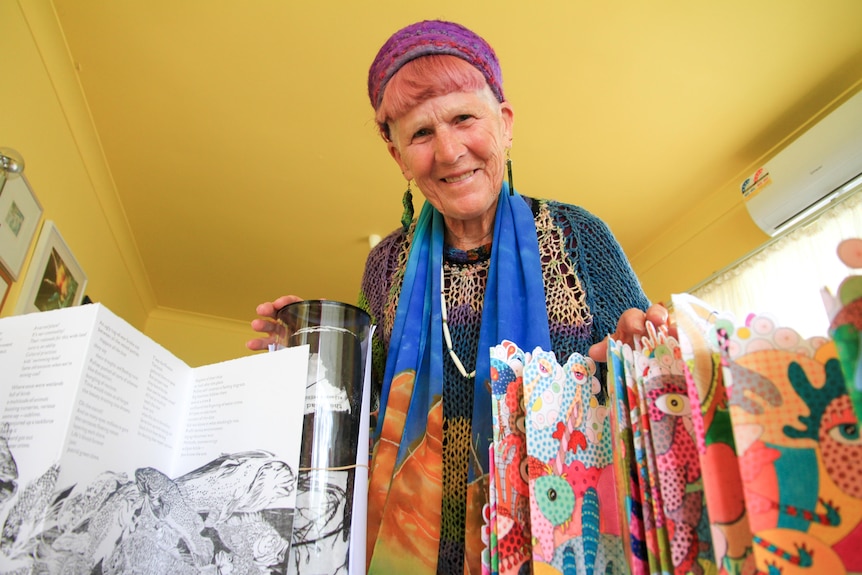 A woman wearing colourful clothes smiles down on the camera which is focussed on the art works before her. 