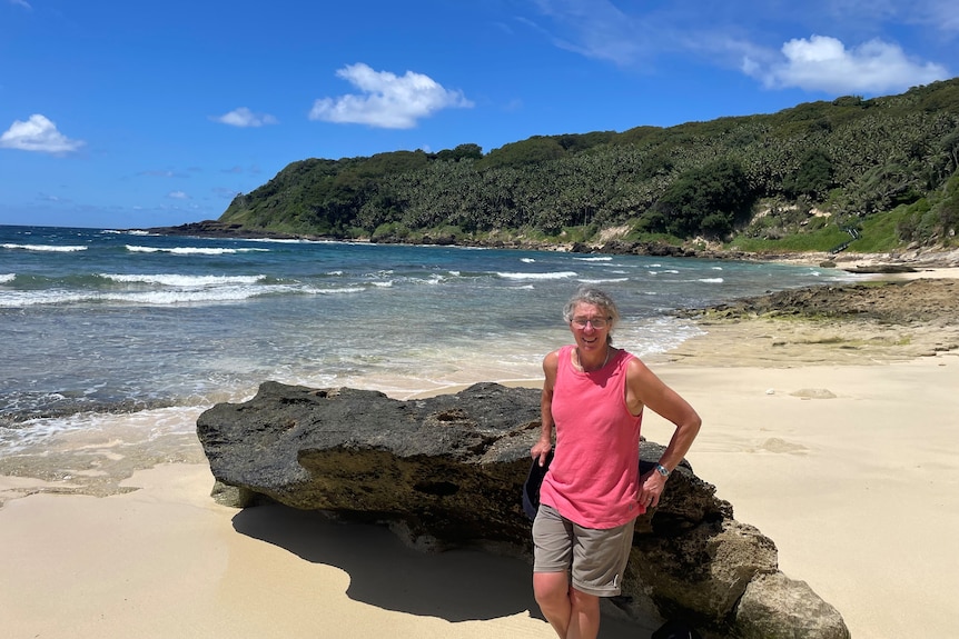 A middle-aged woman stands by a rock on a beach on an island.