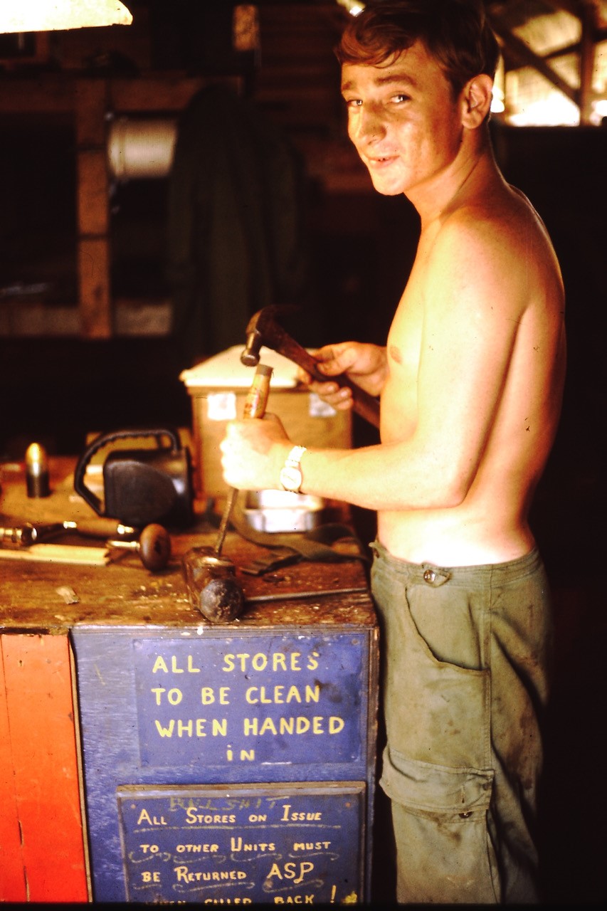 Man, naked to waist, looking at camera and smiling as he hammers a chisel in a store area.