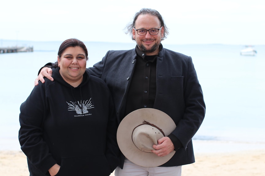 A smiling Indigenous woman and a bespectacled man stand on a beach.