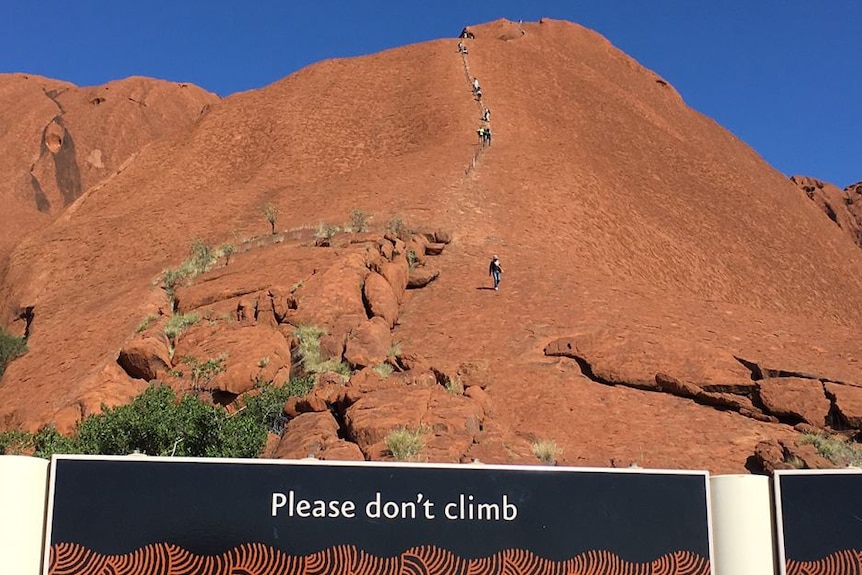 Un panneau devant Uluru, sur lequel marche une file de personnes, qui dit "S'il vous plaît ne grimpez pas".