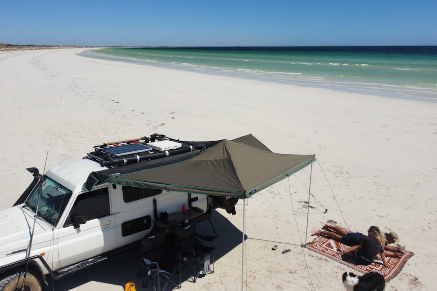 A 4WD with solar panels and an annex parked on beach with a couple lying on towels.