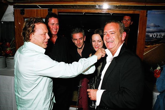 A man in a white shirt adjusts the collar of another man as three people watch on smiling