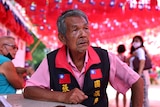 An old man wearing a vest with the Taiwan flag stitched on poses in front of lots of Taiwan flags