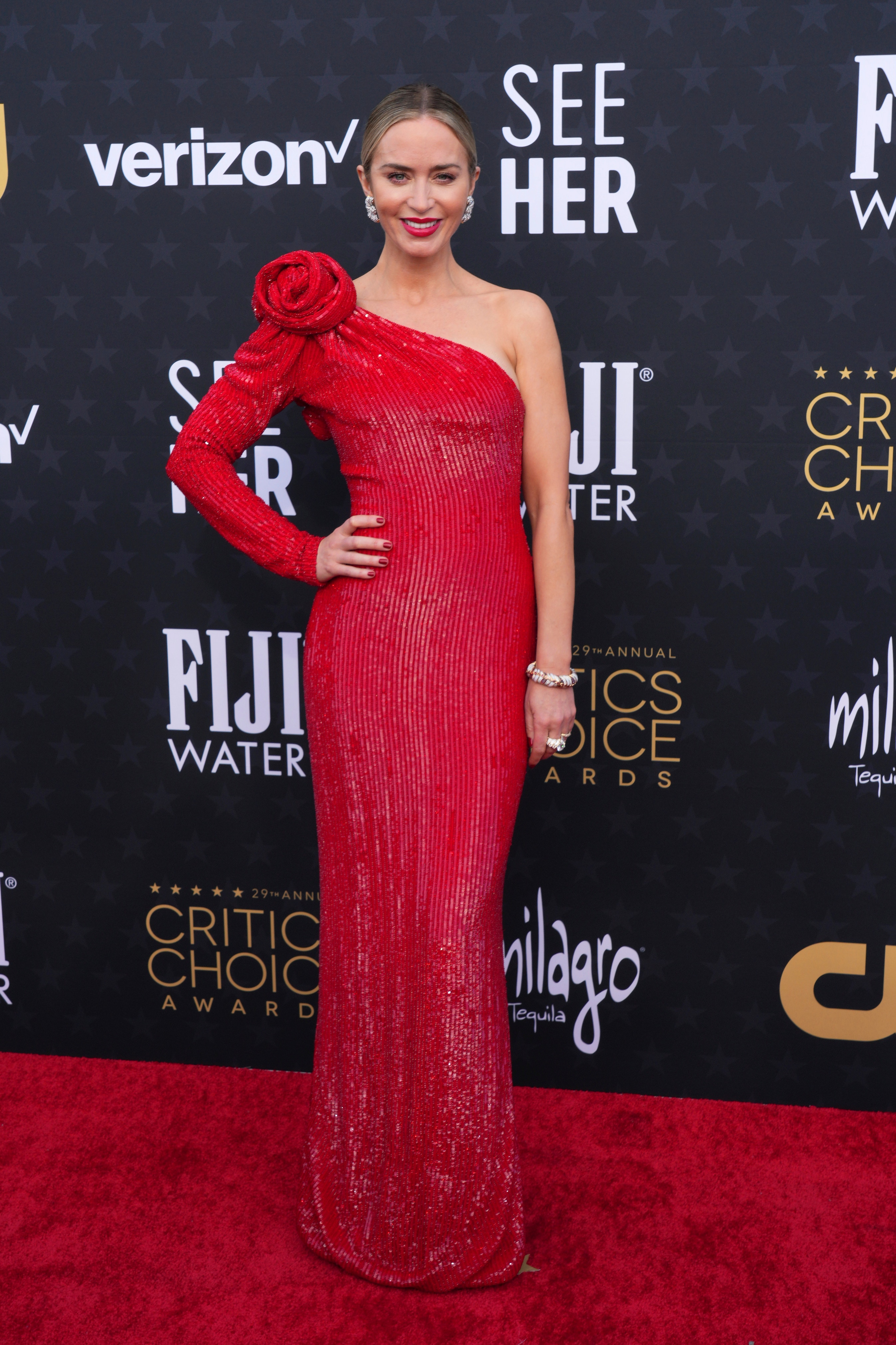 Emily Blunt wearing an off-the-shoulder red gown with a flower on the shoulder with a long sleeve