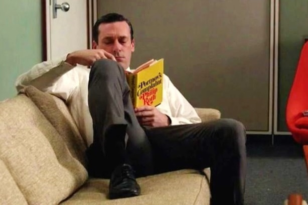 Man sits on couch in an office reading Portnoy's Complaint by Philip Roth