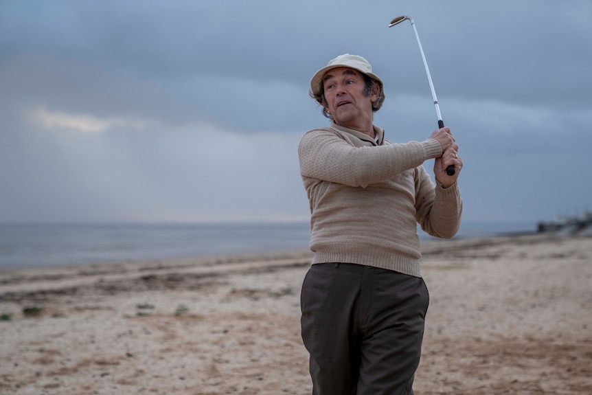 An older man plays golf on a deserted beach on an overcast day, his club raised above his shoulder