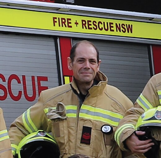 A firefighter poses for a photo with other firefighters. The group are standing in front of a fire truck.