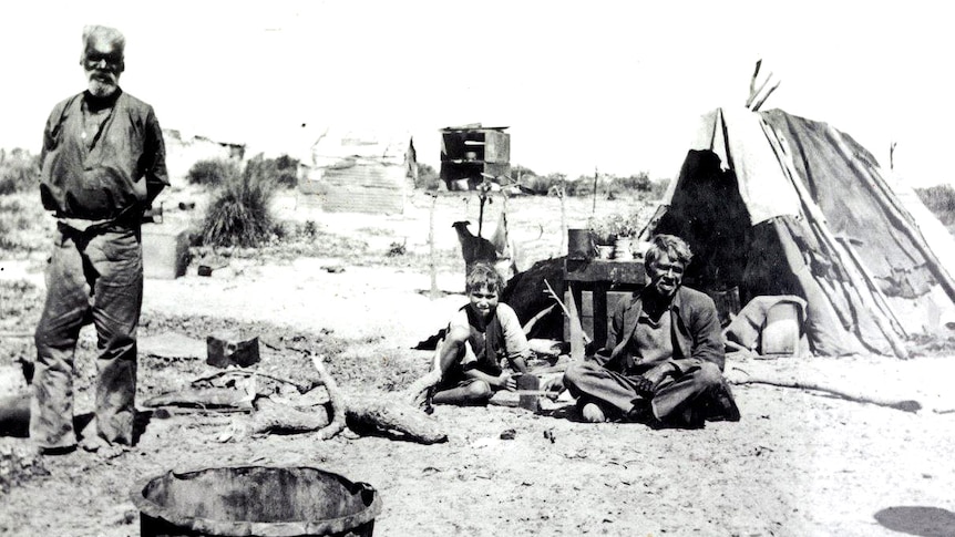 A man standing up, and a man and boy sitting down, in front of a tent.