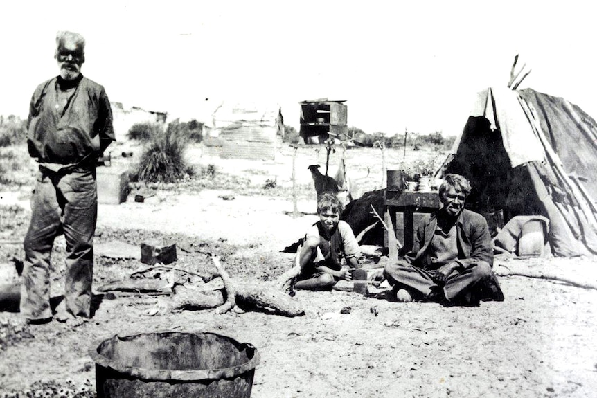 A man standing up, and a man and boy sitting down, in front of a tent.
