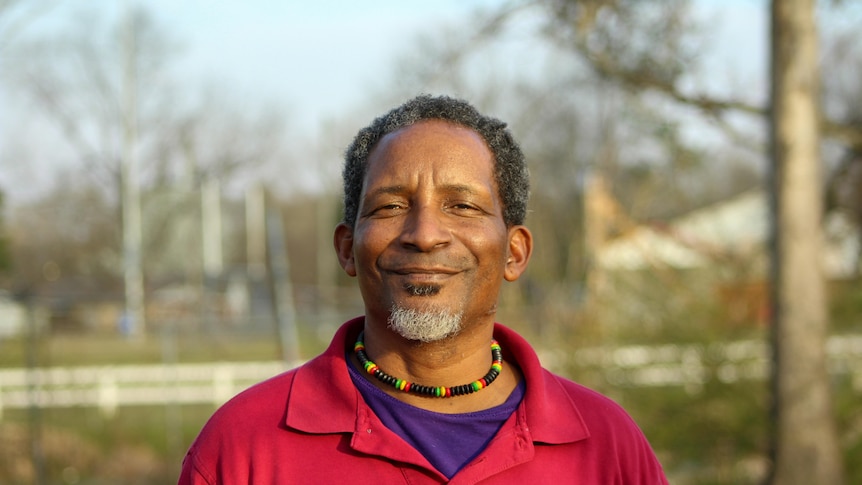 A man in a red shirt smiles at the camera in a park-like setting