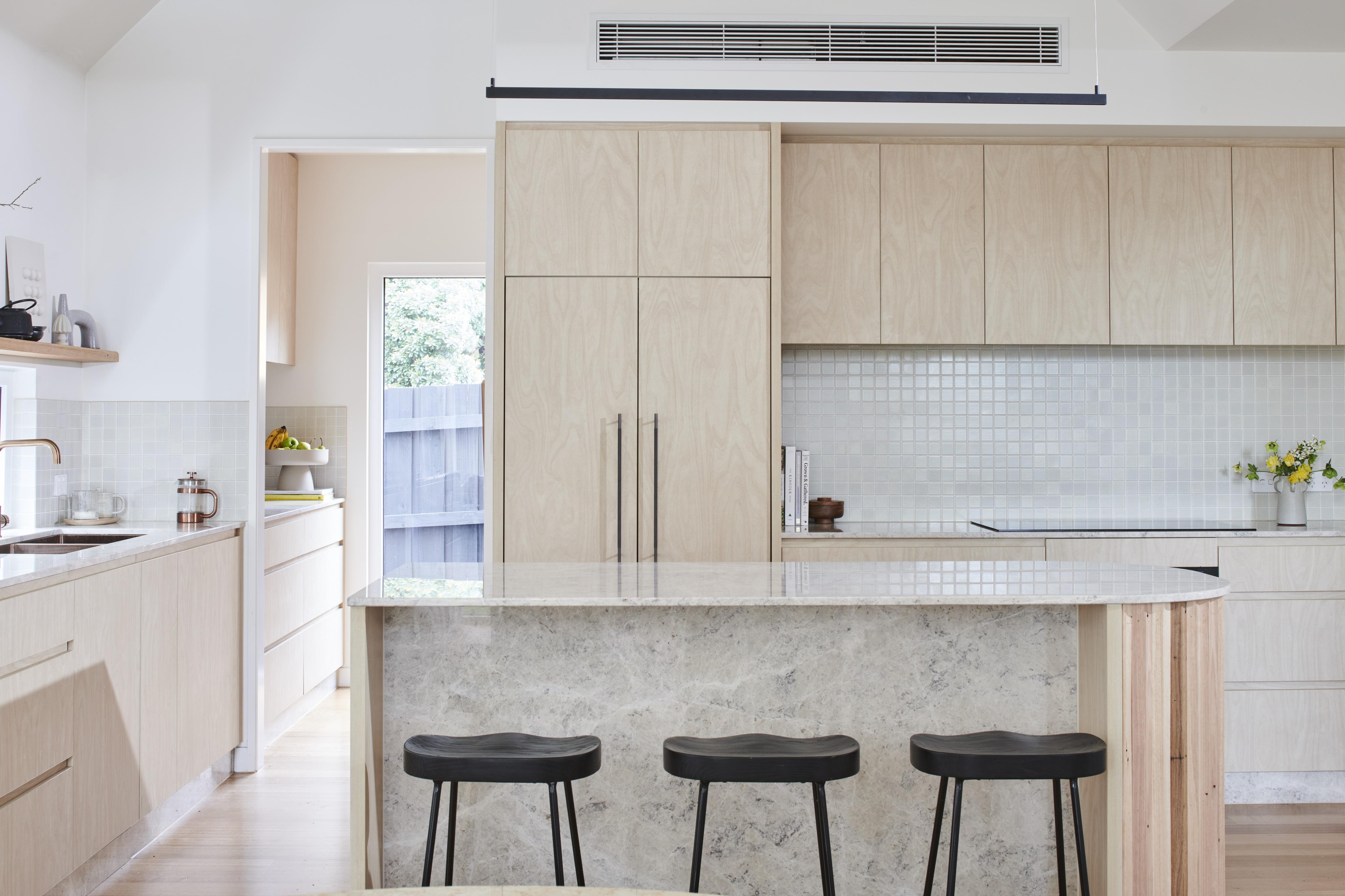 A wide shot of a light filled kitchen with an island bench and bar stools in the middle and light coloured wood cupboards