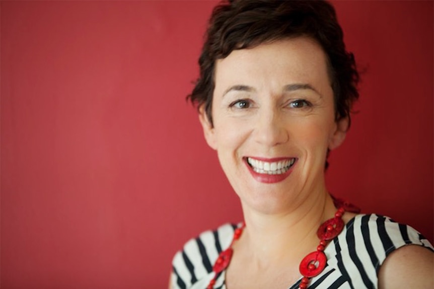 Sarah Macdonald smiling. She has short brown hair and wears a striped shirt and stands in front of a red background.