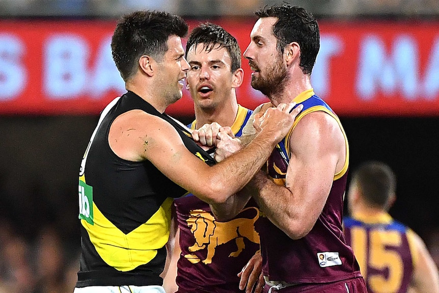 A Richmond AFL player holds the jersey of a Brisbane Lions opponent while they stand facing each other.
