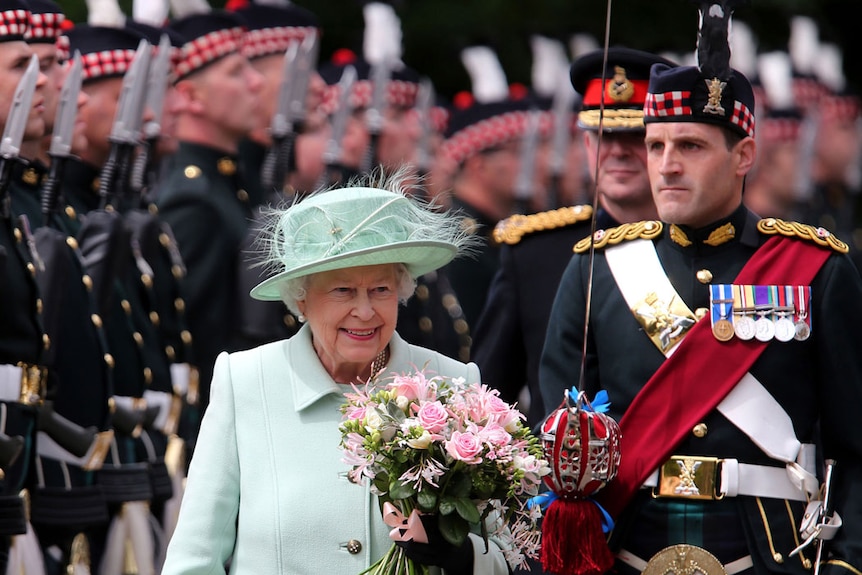 Queen Elizabeth II smiles as she walks past an honour guard of armed soldiers in formal dress uniforms.