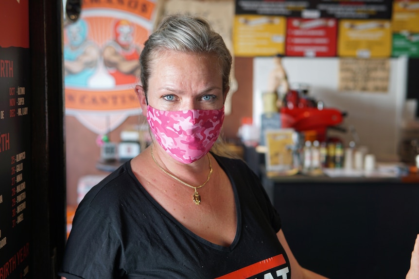 Siri wears a pink mask. She looks into the camera, standing in front of a wall of posters.