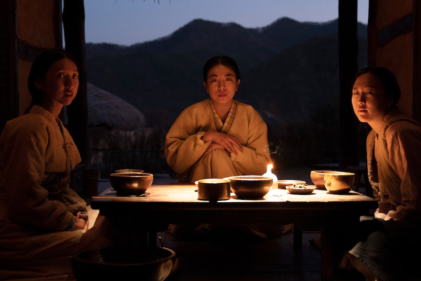 Three women sit at a low table inside a hut in a village. Their pensive faces are lit by candlelight.
