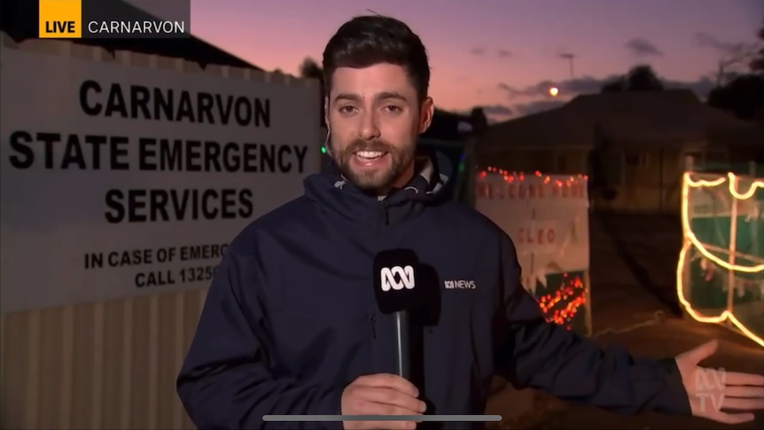 A man wearing a blue ABC jacket and holding a microphone talks on TV in front of fairy lights on an SES base at sunset