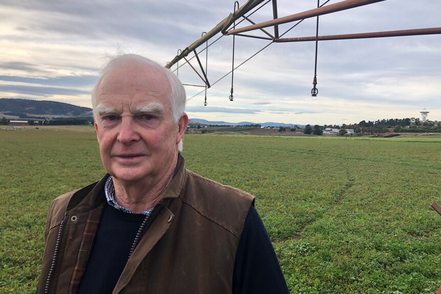 An older man stands in front of an irrigator in a paddock.
