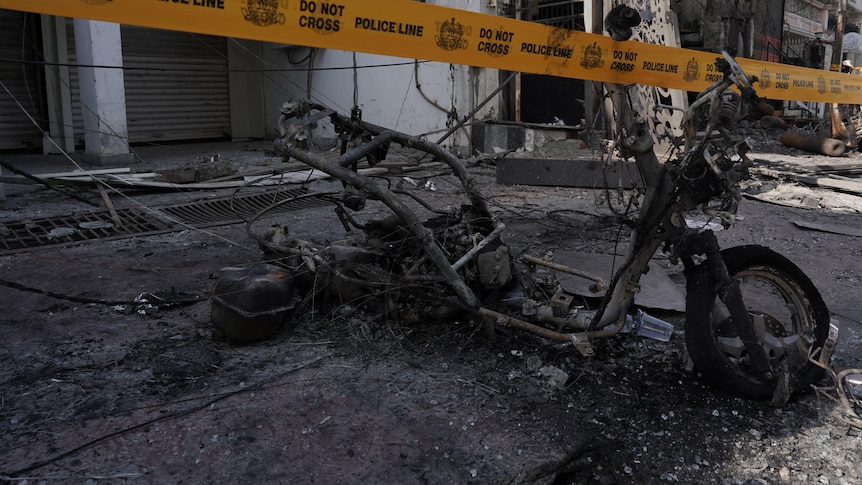 A burned out small bicycle among rubble with yellow police tape in front.