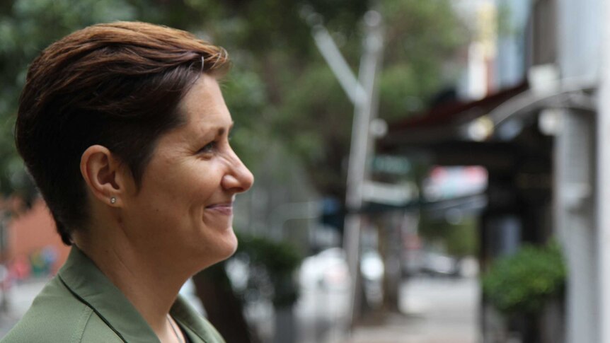 A side shot of a woman smiling. She's standing in front of a quiet, leafy street.