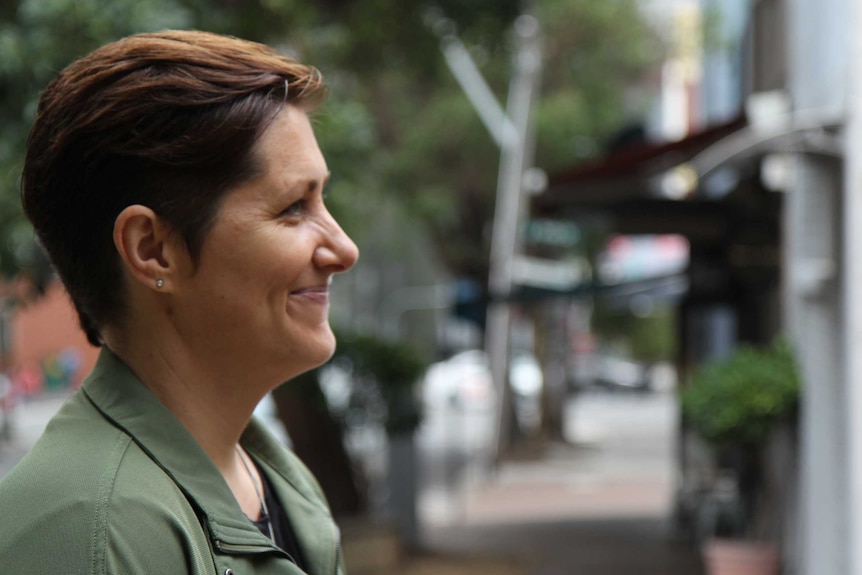 A side shot of a woman smiling. She's standing in front of a quiet, leafy street.