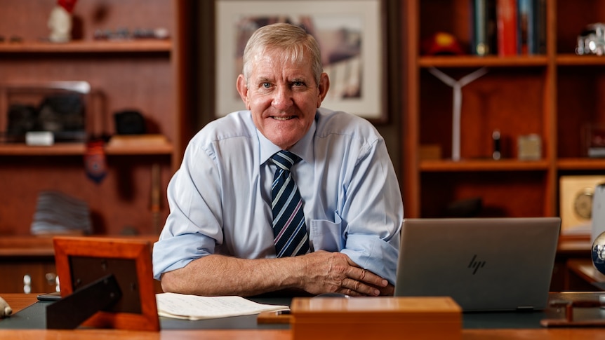 Older man wearing collared shirt and tie sits behind a mahogany desk in an office 