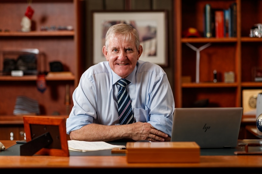 Older man wearing collared shirt and tie sits behind a mahogany desk in an office 