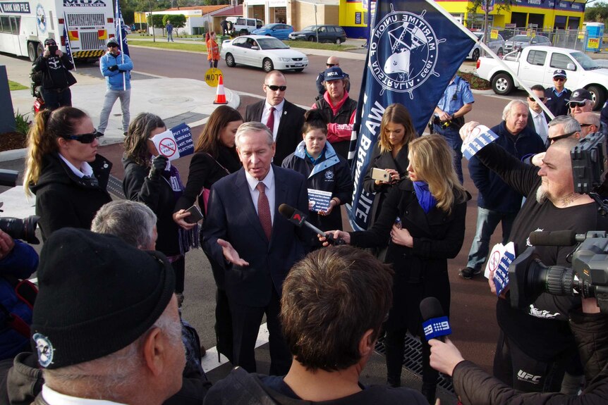 Premier Colin Barnett speaks to reporters as protesting maritime workers look on, with one of them holding a big MUA blue flag.