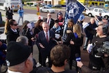 Premier Colin Barnett speaks to reporters as protesting maritime workers look on, with one of them holding a big MUA blue flag.