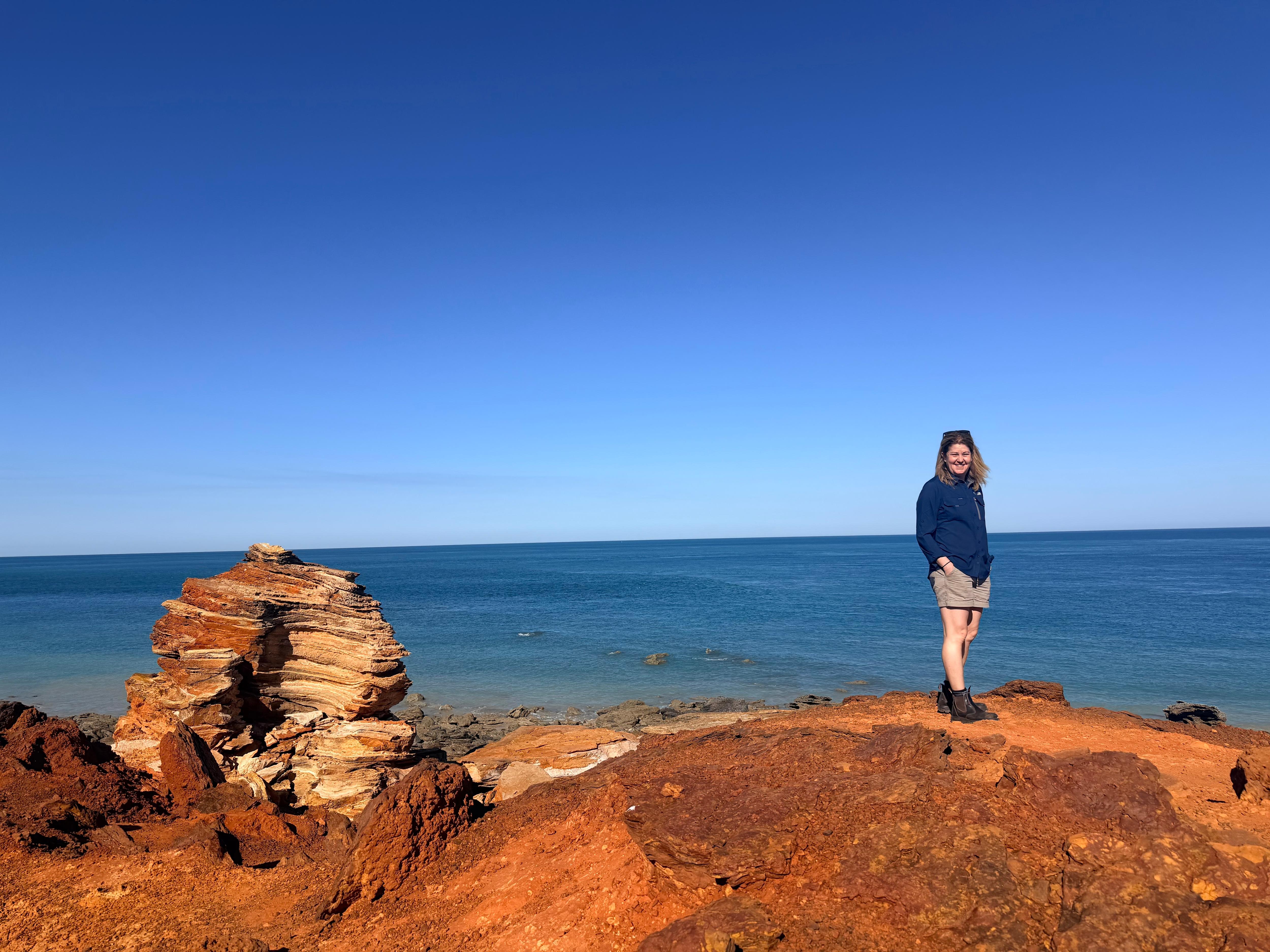 A woman stands on red rock with blue ocean in the background