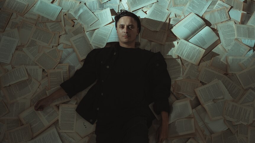 Young man lies on the floor amongst pages of books that have been torn out