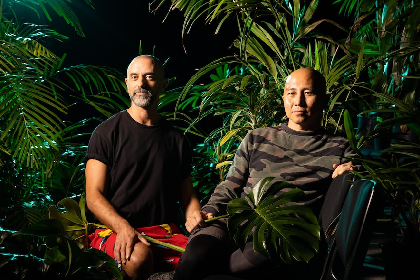 Grey-haired white man in black shirt sits beside Singaporean man with shaved head in army-patterned shirt among plants.