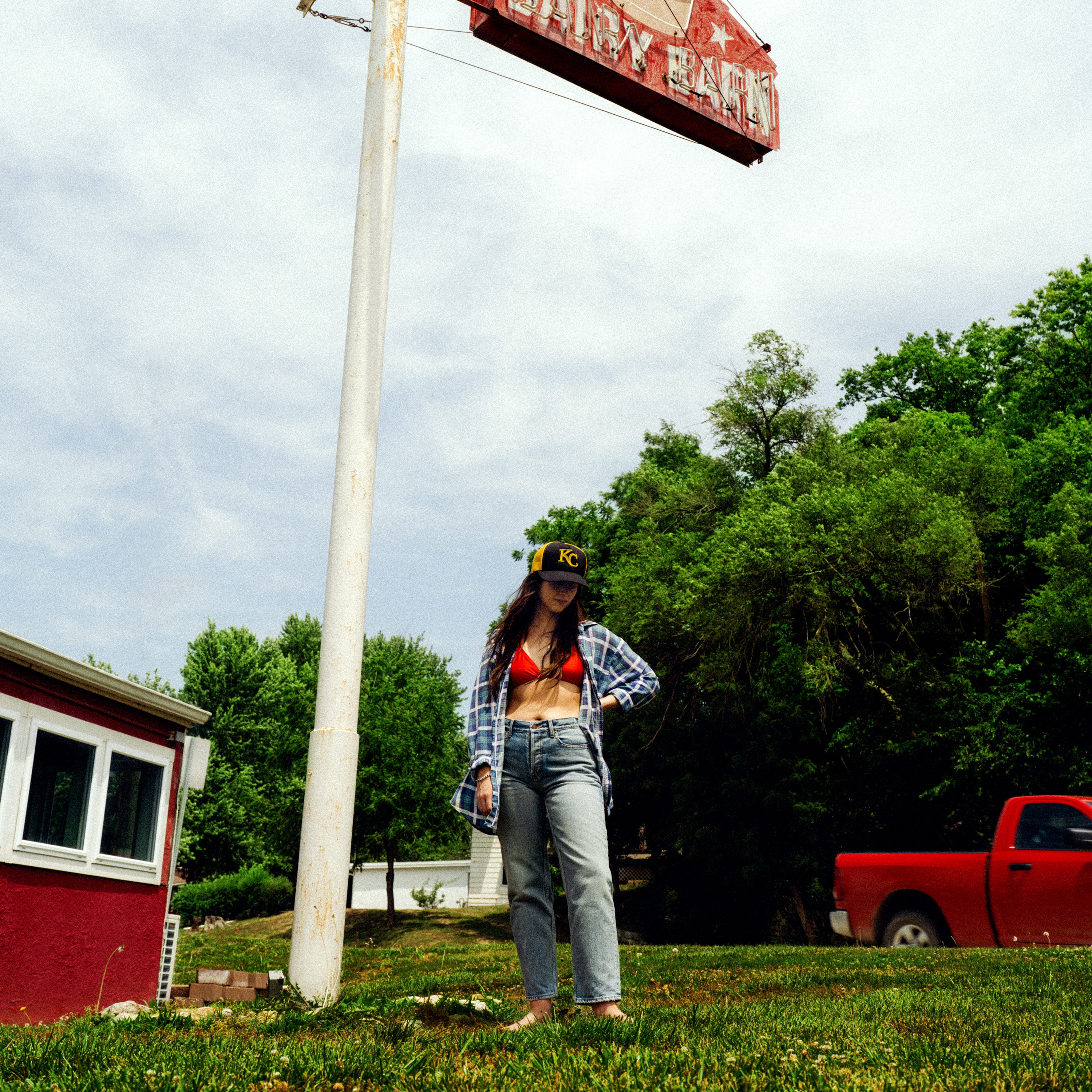 brunette stands in blue jeans, open flannel shirt, red bra and 'KC' trucker cap in field beneath neon sign with red pickup truck
